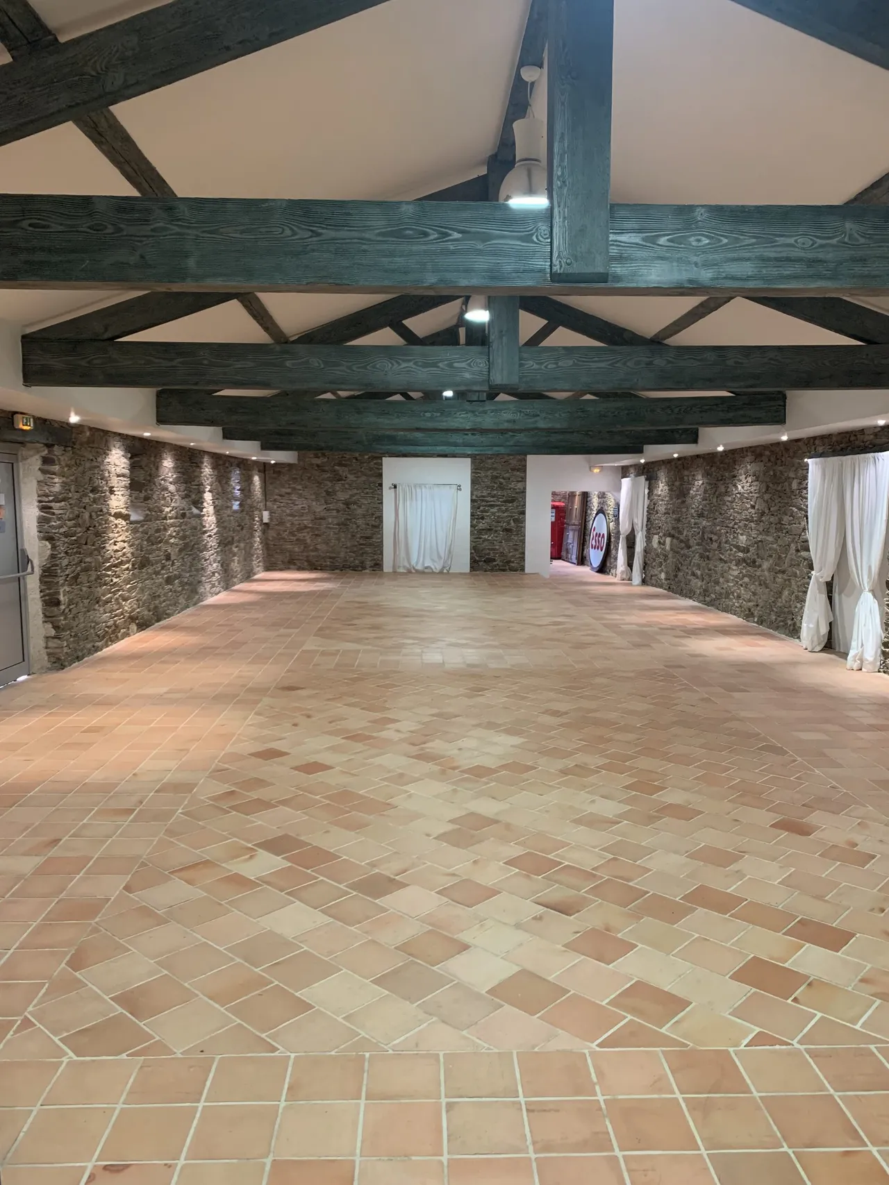 Salle de réception vide : charpente apparente, murs de pierre et sol en tomettes